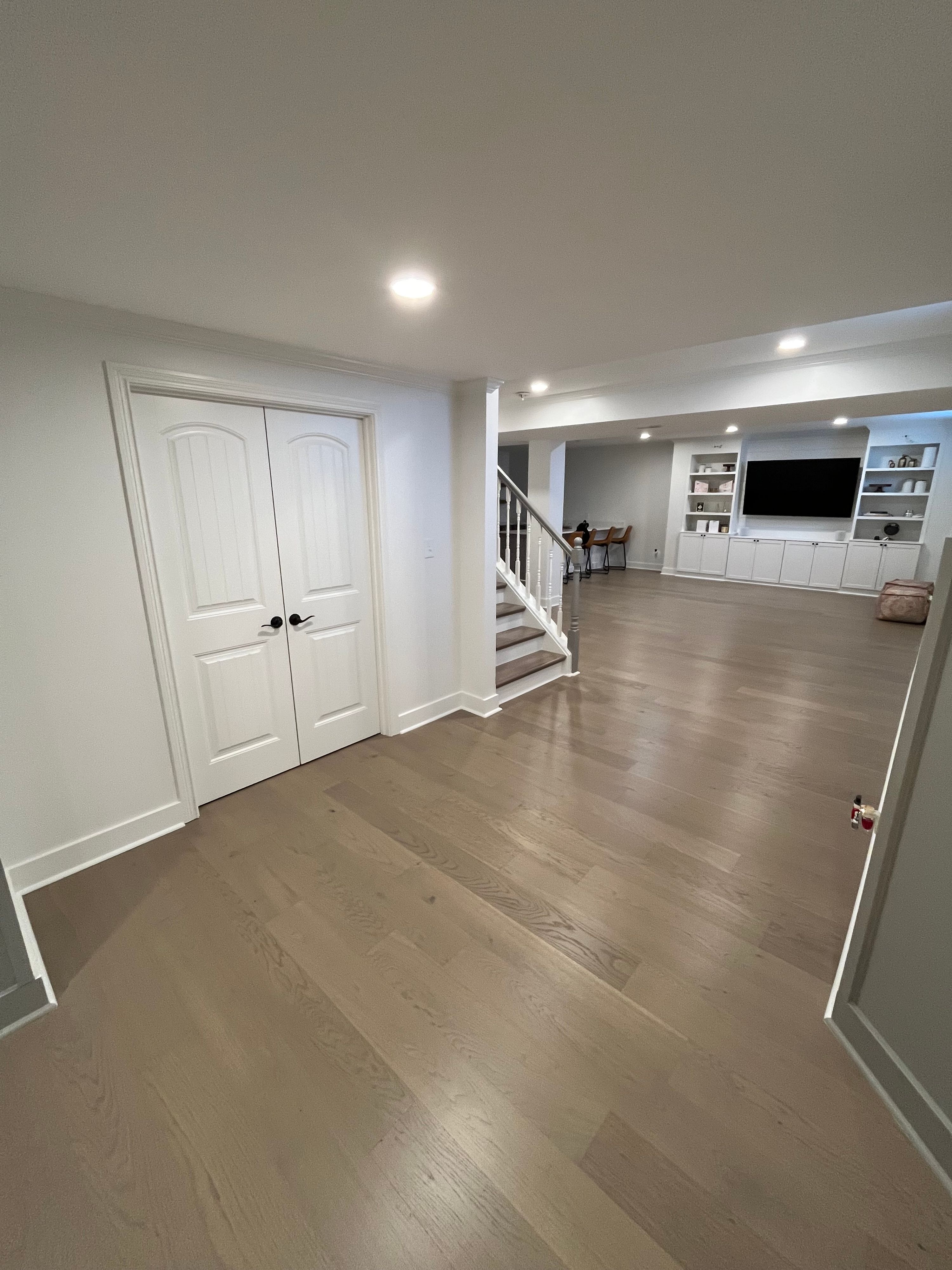 Open layout finished basement with custom entertainment center, white spindle staircase, and wide plank hardwood flooring