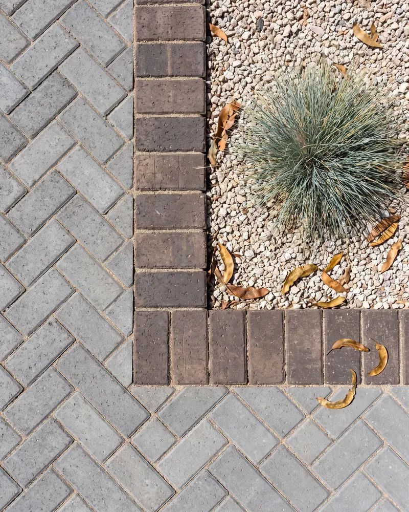 Overhead view of the edge where gray pavers in a herringbone pattern meet a soldier course border and gravel bed