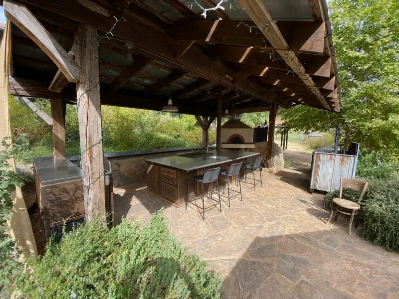 Rustic outdoor kitchen under a timber roof with a long counter, bar stools, and a pizza oven