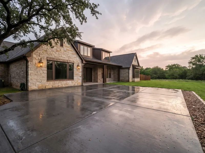 Freshly rained-on broom finished concrete patio draining toward a lawn behind a stone and brick home under clearing clouds