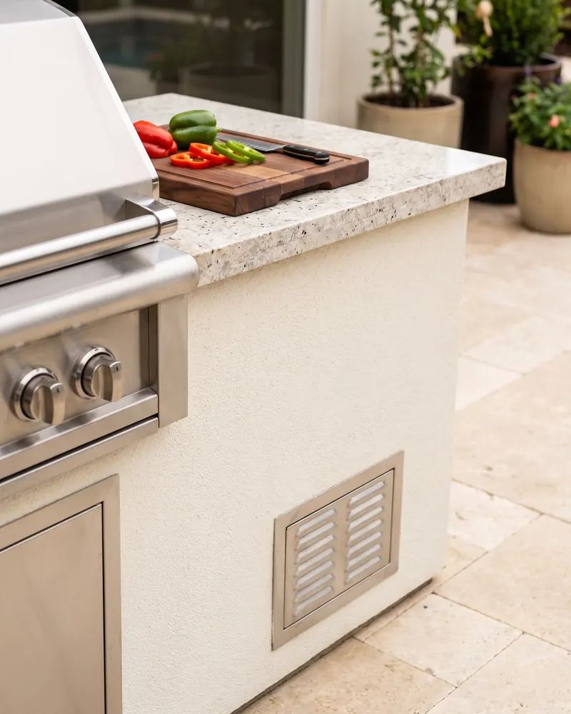 Close view of a light granite counter corner with a stainless grill lid, stucco base and a small vent panel