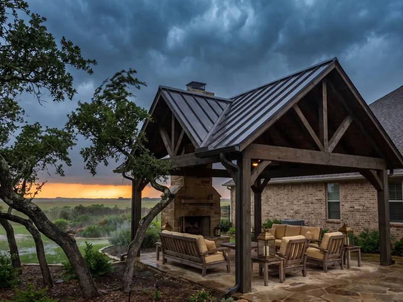 Solid roof pavilion attached to a brick ranch home under a dark spring storm sky, with patio furniture dry beneath it